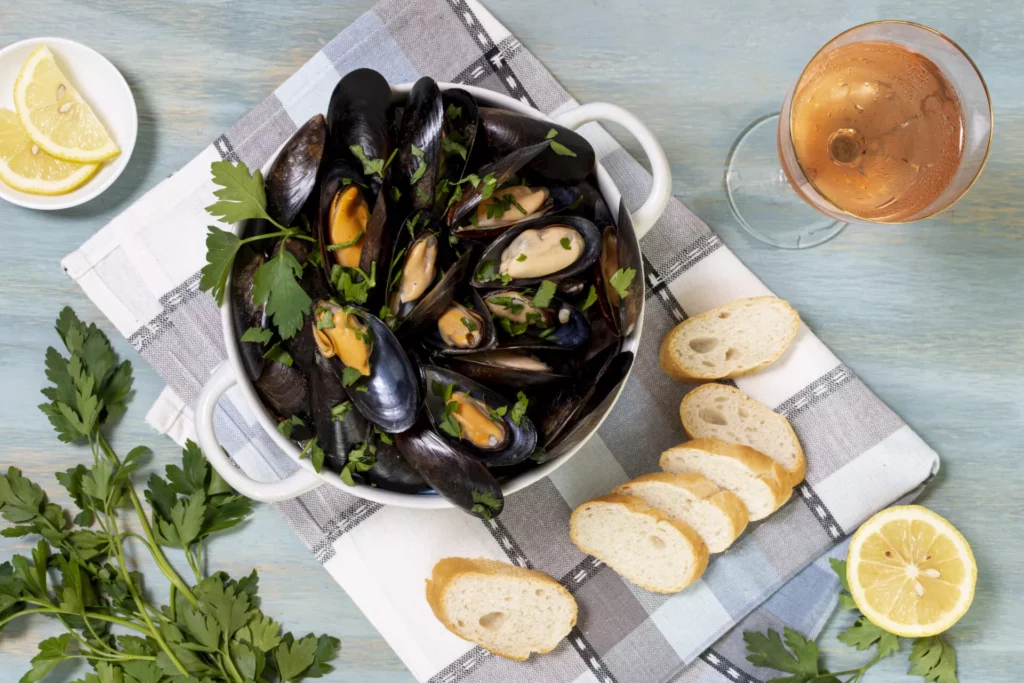 Muscheln essen – Muscheltopf mit Miesmuscheln, Petersilie, Zitronenhälften und Baguettescheiben; daneben Glas Rosé auf hellblauem Holztisch.