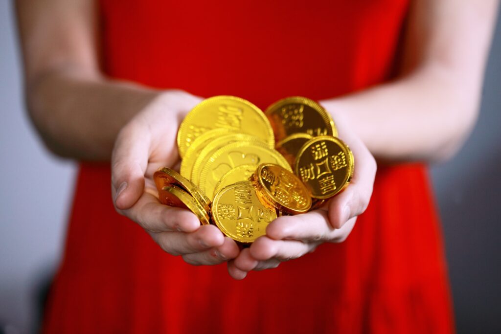 Alt: Hände in rotem Kleid halten mehrere goldene Münzen – Symbolbild für Katja Krasavice Vermögen, Einnahmequellen und finanzielle Erfolge.