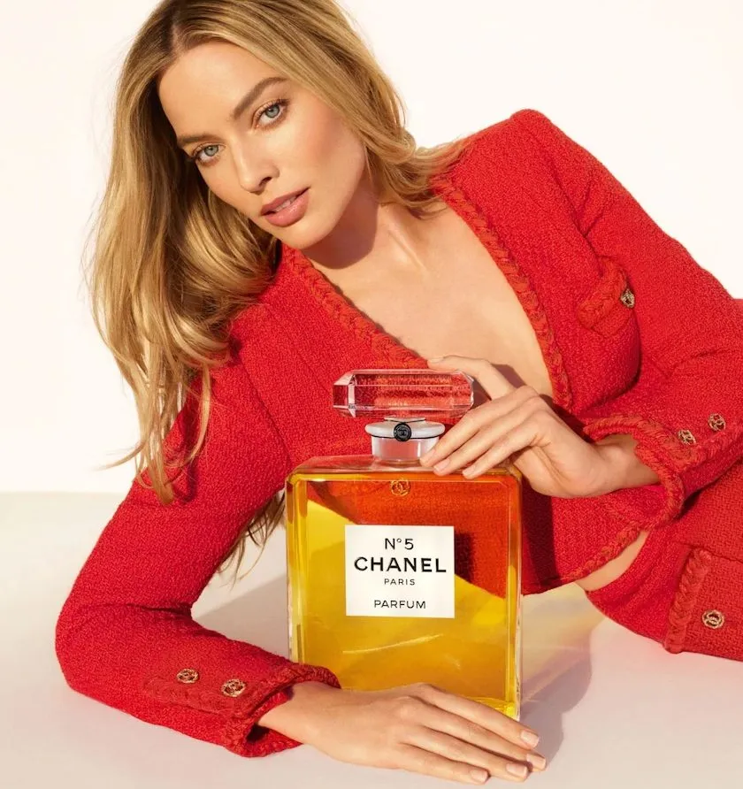 Model in rotem Tweed posiert mit übergroßem Chanel N°5 Parfumflakon; sonniges Licht, luxuriöse Kampagnenästhetik.