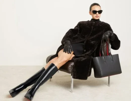 Frau im dunklen Mantel mit hohen schwarzen Louboutin-Stiefeln, Lederhandschuhen und strukturierter Tasche, lässig auf einem Sessel sitzend – moderner Luxuslook 2025.