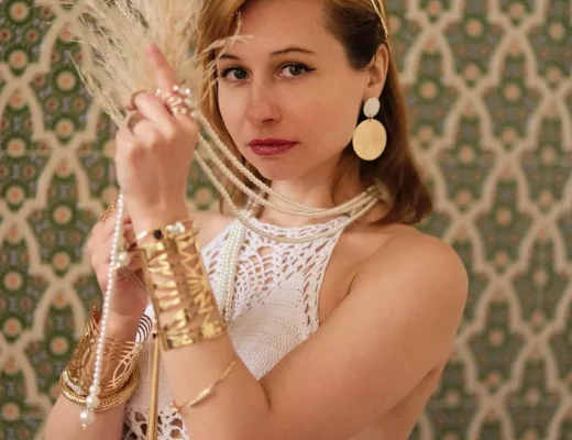 Stilvolle Frau im Boho-Luxus-Look mit weißen Häkel-Details, goldenen Schmuckstücken, auffälligen Ohrringen und glamourösem Lippenstift.