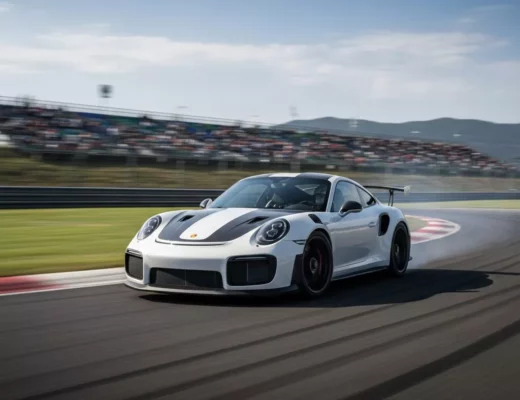 Weißer Porsche 911 GT2 RS Clubsport fährt mit hoher Geschwindigkeit über eine Rennstrecke, Zuschauertribünen im Hintergrund.