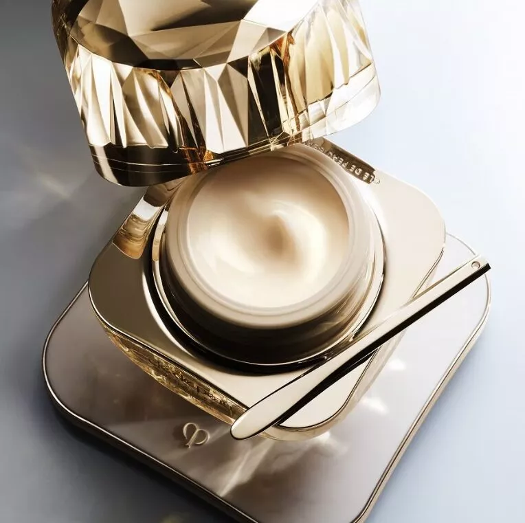 Teuerste Make-up-Marke der Welt – goldfarbener, facettierter Clé-de-Peau-Tiegel mit Spatel auf spiegelnder Ablage, geöffneter Deckel.