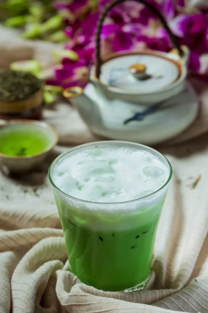 ced Matcha Latte mit dichter Milchschaumhaube im Glas; japanische Teekanne und violette Blüten im Hintergrund.