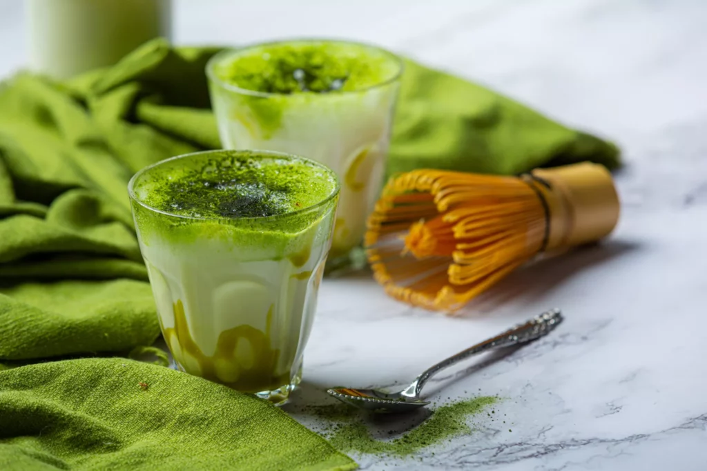 Zwei Gläser Matcha Latte mit grünem Matcha-Shot auf kalter Milch, daneben Bambusbesen (Chasen) und Löffel auf Marmoruntergrund.