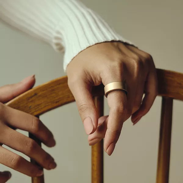 Hand mit goldenem Ultrahuman Ring AIR, getragen als luxuriöses Schmuckstück auf einem Holzstuhl