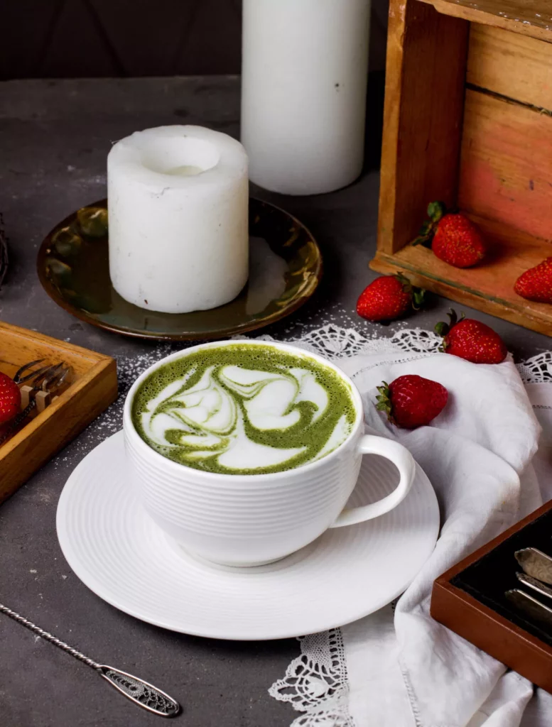 Heißer Matcha Latte mit marmorierter Latte-Art in weißer Rillentasse; Erdbeeren und Kerzen als Food-Styling.
