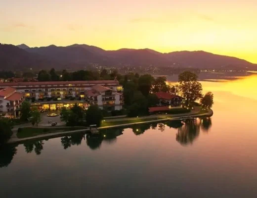 Althoff Seehotel Überfahrt am Tegernsee – Luftaufnahme bei Sonnenuntergang mit Halbinsel, ruhigem Wasser und Alpen-Silhouette in Rottach-Egern, Bayern.
