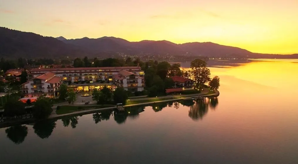 Althoff Seehotel Überfahrt am Tegernsee – Luftaufnahme bei Sonnenuntergang mit Halbinsel, ruhigem Wasser und Alpen-Silhouette in Rottach-Egern, Bayern.