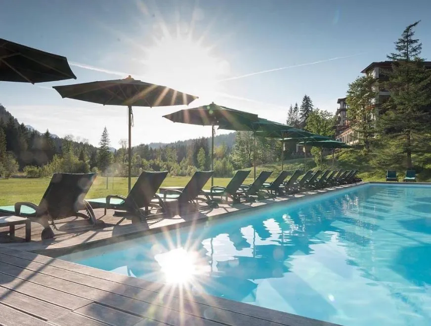 Schloss Elmau Luxury Spa Retreat – Außenpool mit Liegestühlen und Sonnenschirmen vor Alpenkulisse im Morgenlicht, Bayern, Deutschland.