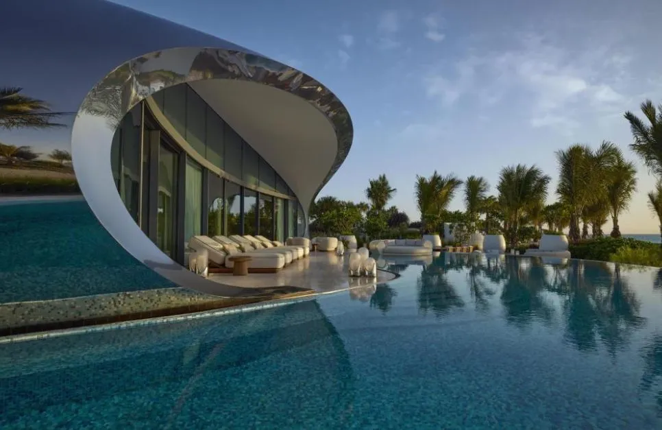 Organisch geschwungene Villa des Shebara Resort mit offenem Lounge-Bereich und großem, spiegelglattem Pool, umgeben von Palmen bei spätem Nachmittagslicht.