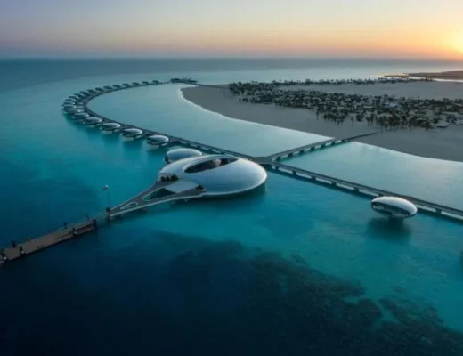 Luftaufnahme des Shebara Resort auf Shebara Island mit ringförmig angeordneten Überwasser-Villen, futuristischem Zentralgebäude und Stegen bei Sonnenuntergang über dem Roten Meer.