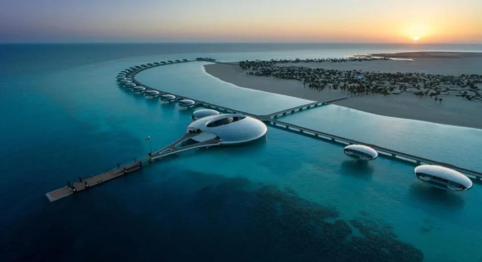 Luftaufnahme des Shebara Resort auf Shebara Island mit ringförmig angeordneten Überwasser-Villen, futuristischem Zentralgebäude und Stegen bei Sonnenuntergang über dem Roten Meer.