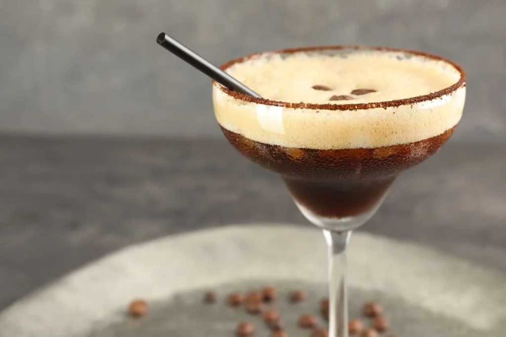 Espresso Martini in einer Coupette mit feiner Crema und Schokoladenrand auf grauer Oberfläche, Nahaufnahme mit Kaffeebohnen im Hintergrund.