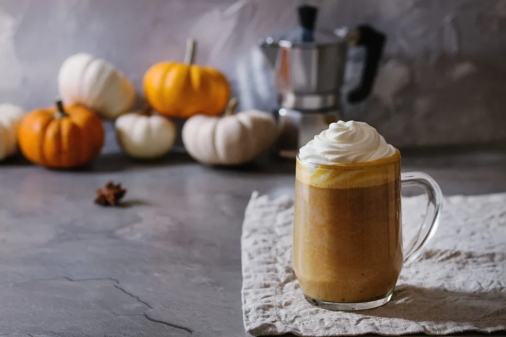 Pumpkin Spice Latte im Glasbecher mit Sahnetopping, im Hintergrund kleine Zierkürbisse und Espressokocher auf Steinuntergrund