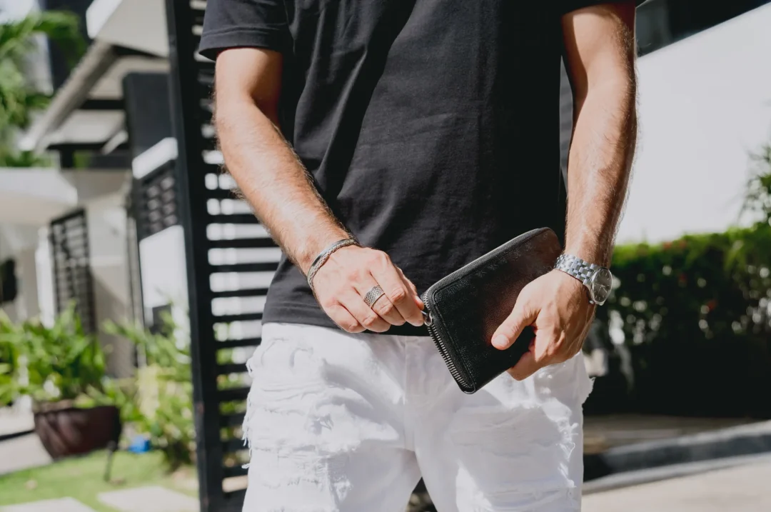 Mann in schwarzem T-Shirt und weißen Jeans hält eine schwarze Leder-Clutch mit Reißverschluss im Freien.