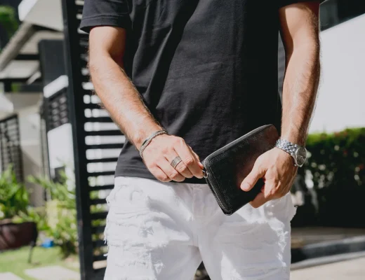 Mann in schwarzem T-Shirt und weißen Jeans hält eine schwarze Leder-Clutch mit Reißverschluss im Freien.