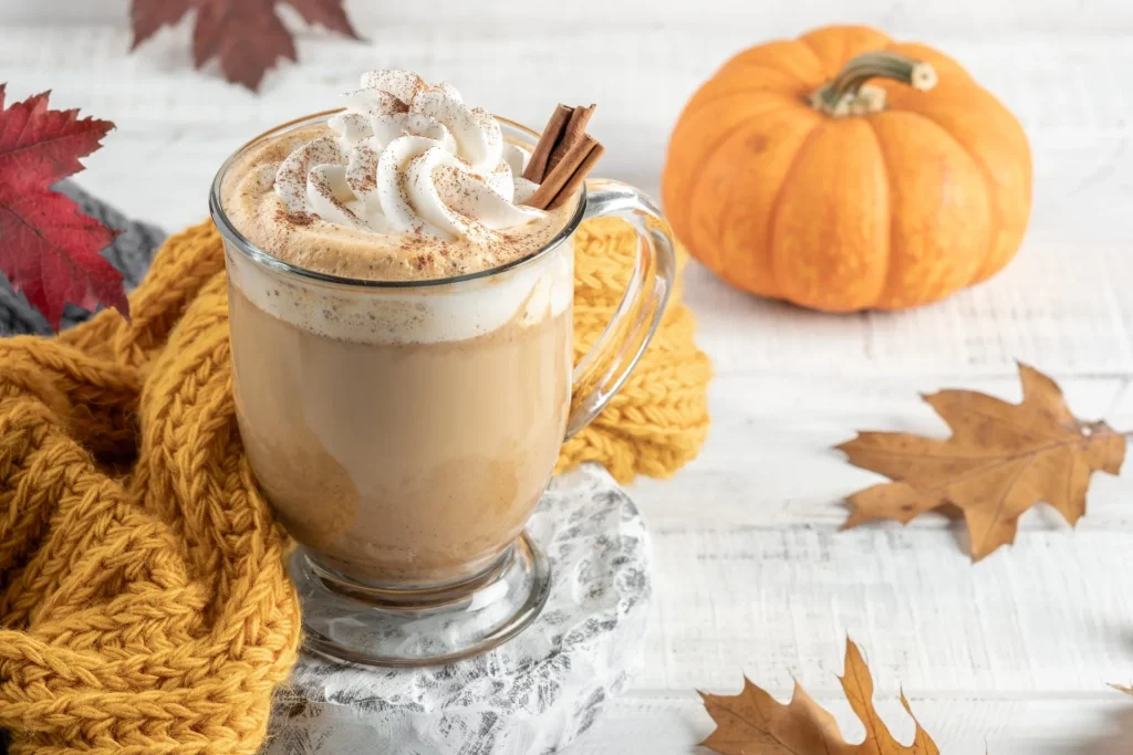 Pumpkin Spice Latte im Glas mit Sahnehaube, Zimtstange und Zimtpuder, dekoriert mit kleinem Kürbis und Herbstblättern auf hellem Holz