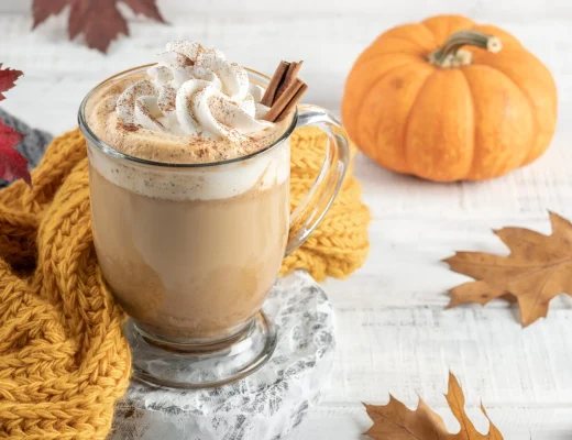 Pumpkin Spice Latte im Glas mit Sahnehaube, Zimtstange und Zimtpuder, dekoriert mit kleinem Kürbis und Herbstblättern auf hellem Holz