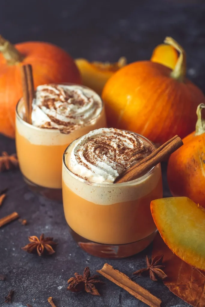 Zwei Gläser Pumpkin Spice Latte mit Sahnewirbel und Zimtstangen, umgeben von Hokkaido-Kürbissen, Sternanis und Zimtrinde auf dunklem Stein