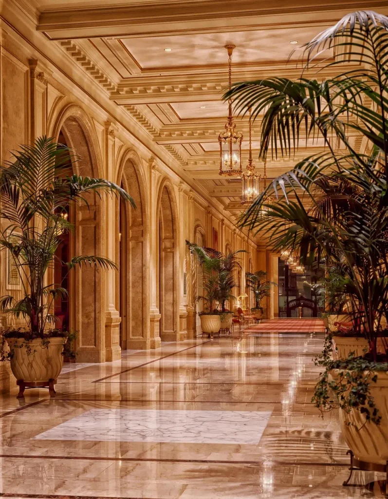 Grandhotel-Lobby mit Marmorboden, klassizistischen Arkaden, goldenen Laternenleuchten und großen Kübelpalmen – repräsentativer Hotelflur bei Abendbeleuchtung.