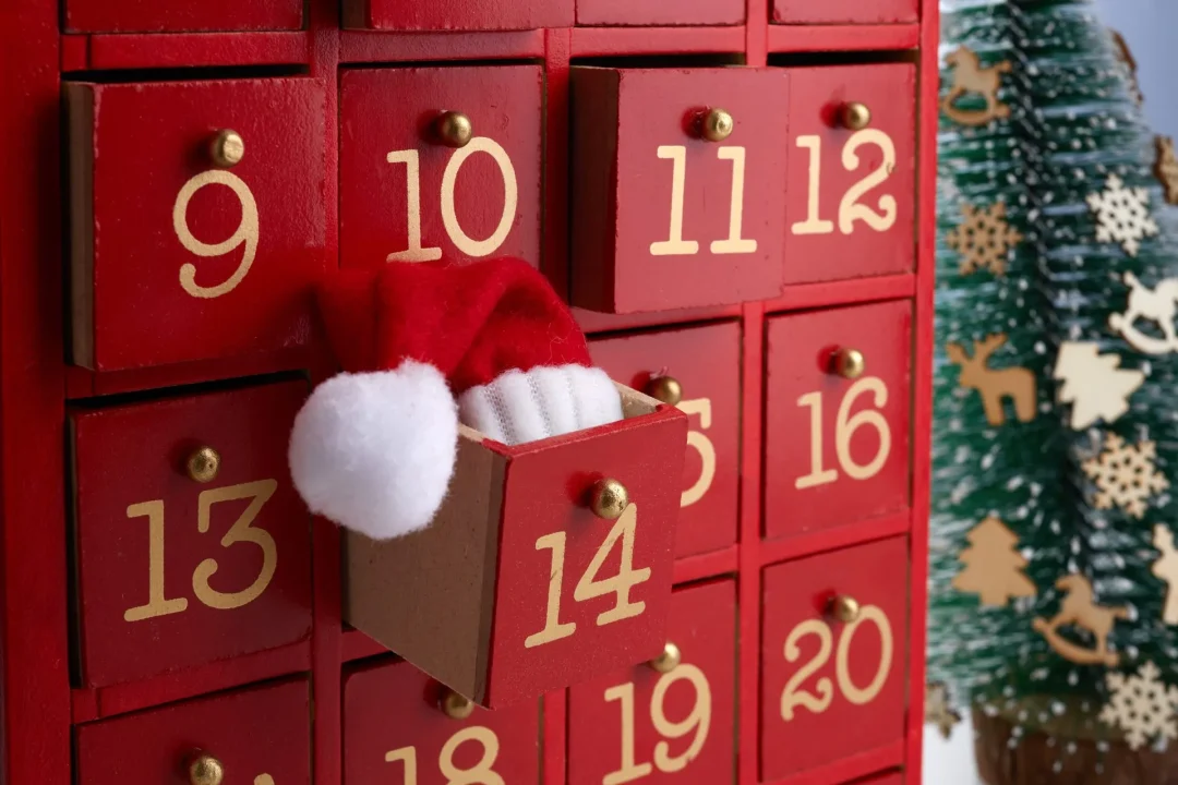 Roter Holz-Adventskalender mit nummerierten Schubladen; hinter Türchen 14 ragt eine kleine Nikolausmütze hervor – stimmungsvolle Szene für Adventskalender für Frauen 2025.