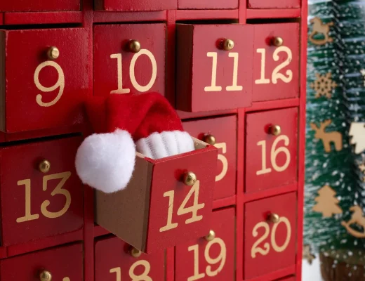 Roter Holz-Adventskalender mit nummerierten Schubladen; hinter Türchen 14 ragt eine kleine Nikolausmütze hervor – stimmungsvolle Szene für Adventskalender für Frauen 2025.