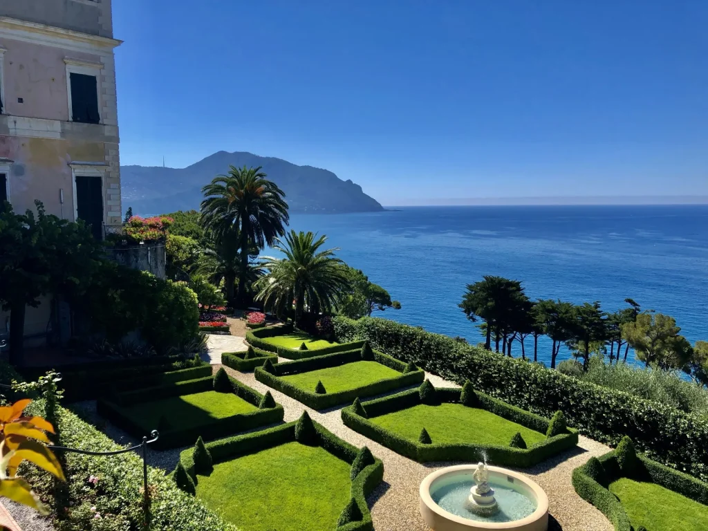 Exklusive Immobilien in Italien – historischer Palastgarten mit Buchsgeometrien, Palmen und Meerblick über die ligurische Küste bei klarem Himmel.