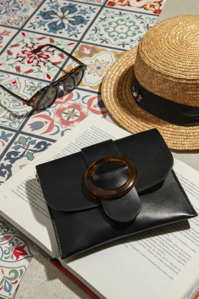 Schwarze Leder-Clutch mit runder Schließe auf offenem Buch, daneben Sonnenhut und Sonnenbrille auf mediterranen Fliesen