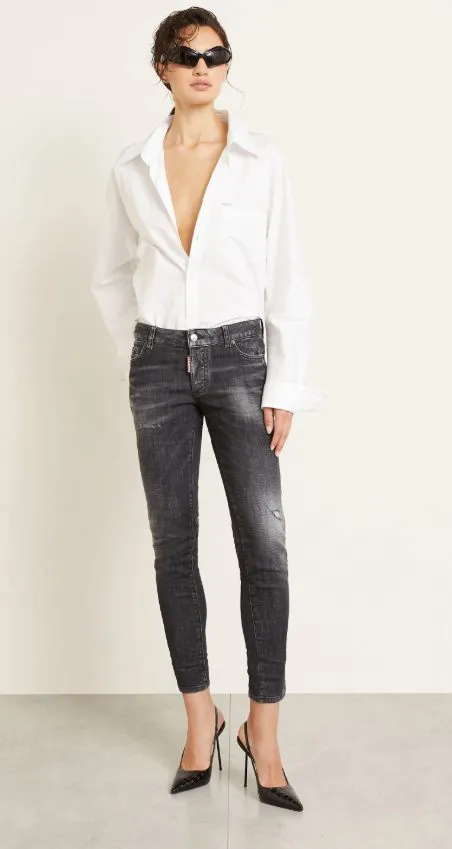 Dame in ausgewaschener grauer Skinny Jeans, offen getragenem weißen Hemd und schwarzen High Heels, stehend als moderner Skinny-Jeans-Look 2025.