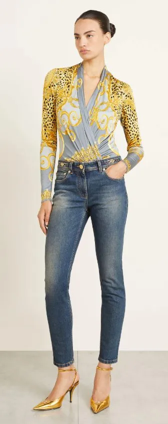 Dame in mittelblauer Skinny Jeans, gemustertem Wickel-Body in Grau-Gold und goldenen Slingback-Heels, stehend als Beispiel für einen eleganten Skinny-Jeans-Look 2025.