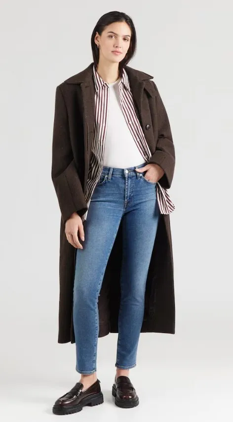 Dame in mittelblauer Skinny Jeans, weißem Shirt, gestreiftem Hemd und langem dunkelbraunem Mantel, kombiniert mit Loafern – moderner Winterlook 2025.