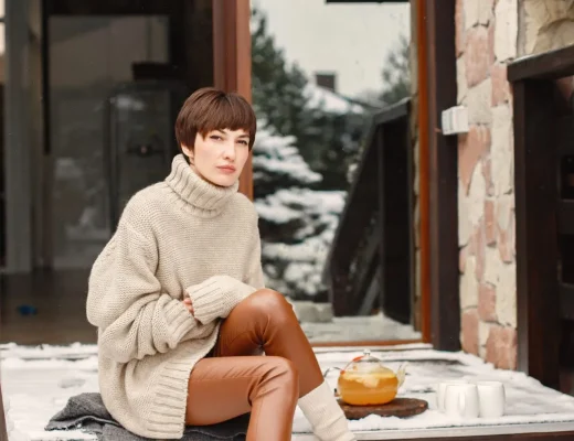 Frau trägt moderne Ugg Boots mit dicken Socken, einem oversized Rollkragenpullover und brauner Lederleggings auf schneebedeckten Holzstufen vor einem Haus.