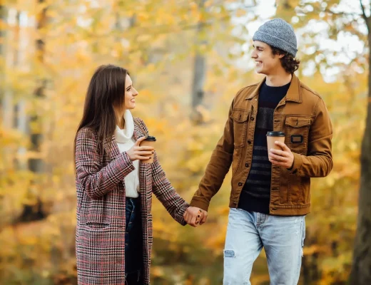 Paar hält Händchen bei einem Herbstspaziergang und trinkt Kaffee – symbolische Darstellung der Cuffing Season