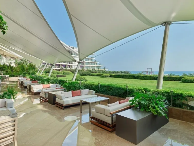 &Uuml;berdachter Lounge-Bereich im Maxx Royal Belek mit eleganten Sitzm&ouml;beln, Blick auf Gartenanlage und Mittelmeer.