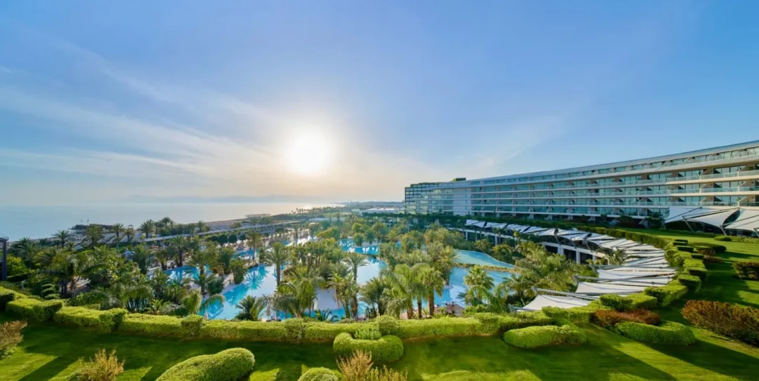 Weitläufige Pool- und Gartenlandschaft des Maxx Royal Belek Golf Resort mit Blick auf das Hauptgebäude und das Mittelmeer bei Sonnenlicht.