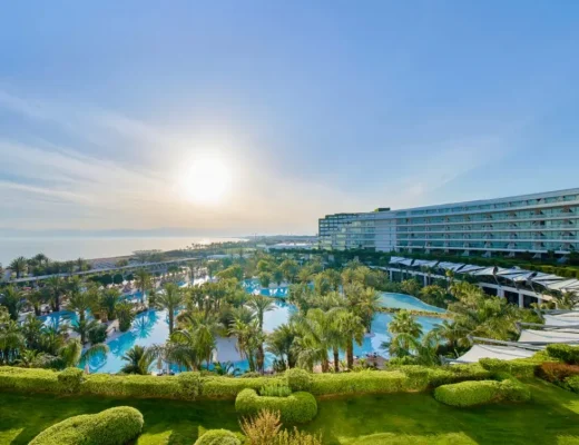 Weitläufige Pool- und Gartenlandschaft des Maxx Royal Belek Golf Resort mit Blick auf das Hauptgebäude und das Mittelmeer bei Sonnenlicht.
