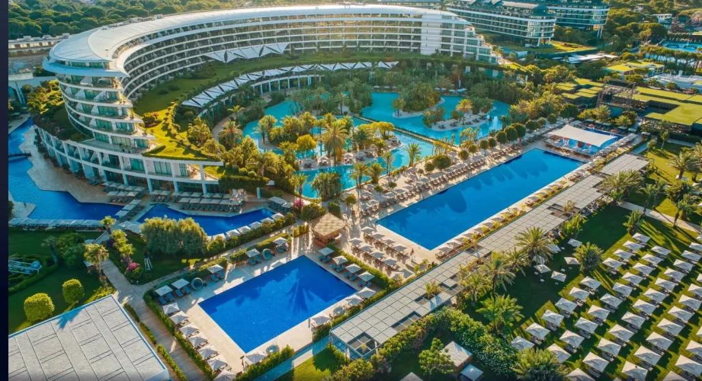 Luftaufnahme der Pool- und Gartenanlage des Maxx Royal Belek Golf Resort mit Hauptgeb&auml;ude und Palmenlandschaft.