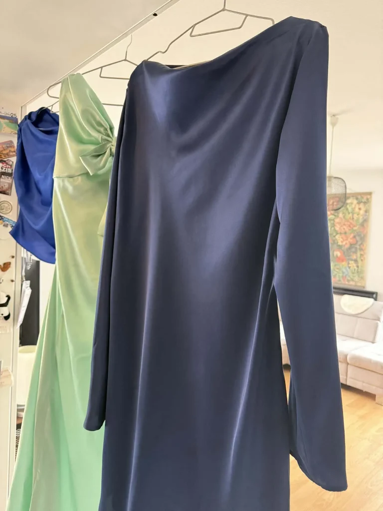 Seidenbluse und Seidenkleider von Melisande Spenger in Blau- und Grüntönen auf Bügeln.