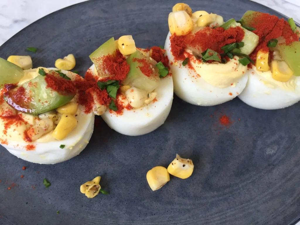 Deviled Eggs mit cremiger Eigelbfüllung, Paprikapulver, Mais, Kräutern und eingelegtem Gemüse, modern angerichtet auf dunklem Teller