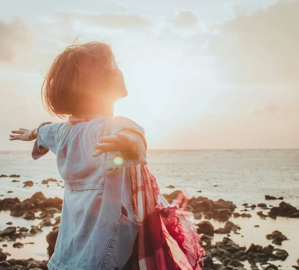 Frau breitet am Meer bei Sonnenaufgang die Arme aus – Symbol für Longevity, Gesundheit, Achtsamkeit und ein langes, aktives Leben.