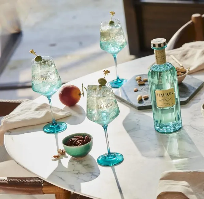 Drei Gläser Italicus Spritz mit Eis und grünen Oliven neben einer Flasche Italicus Rosolio di Bergamotto auf einem Marmortisch.