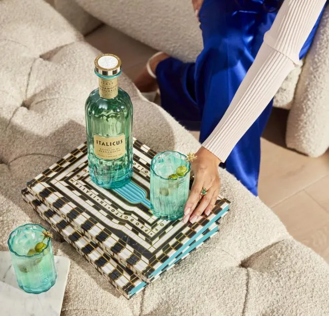 Flasche Italicus Rosolio di Bergamotto neben zwei Gläsern Italicus Spritz mit Eis und grünen Oliven auf einem dekorierten Couchtisch.