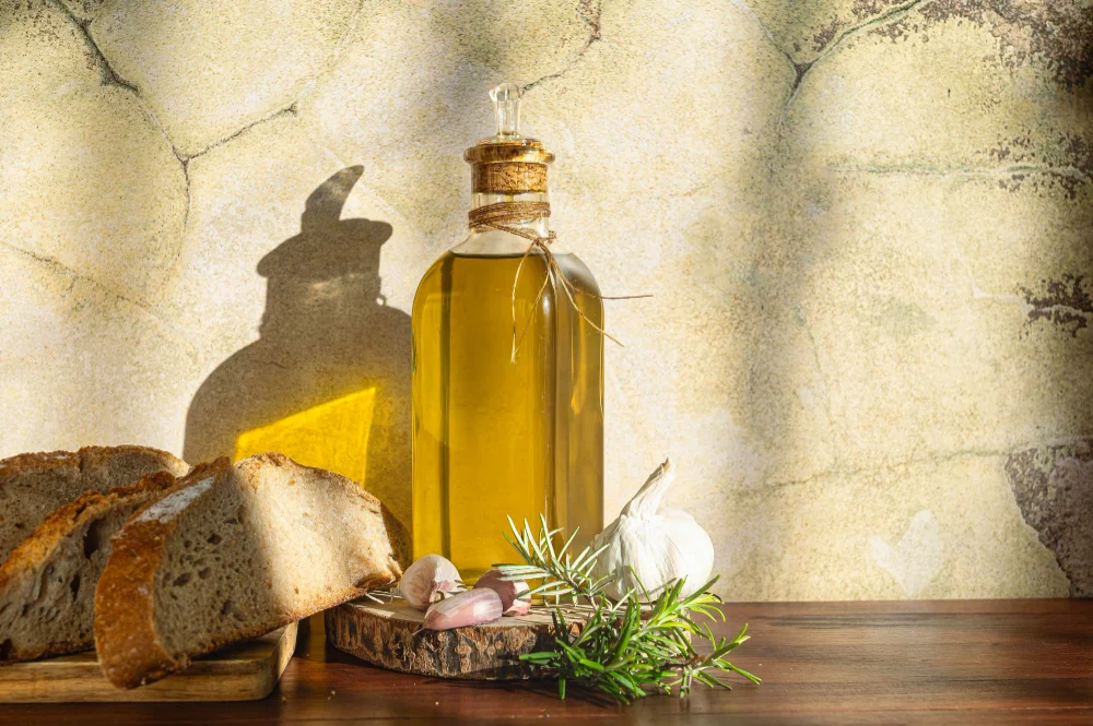Flasche mit goldenem Olivenöl neben rustikalem Brot, Knoblauch und Rosmarin auf einem Holztisch