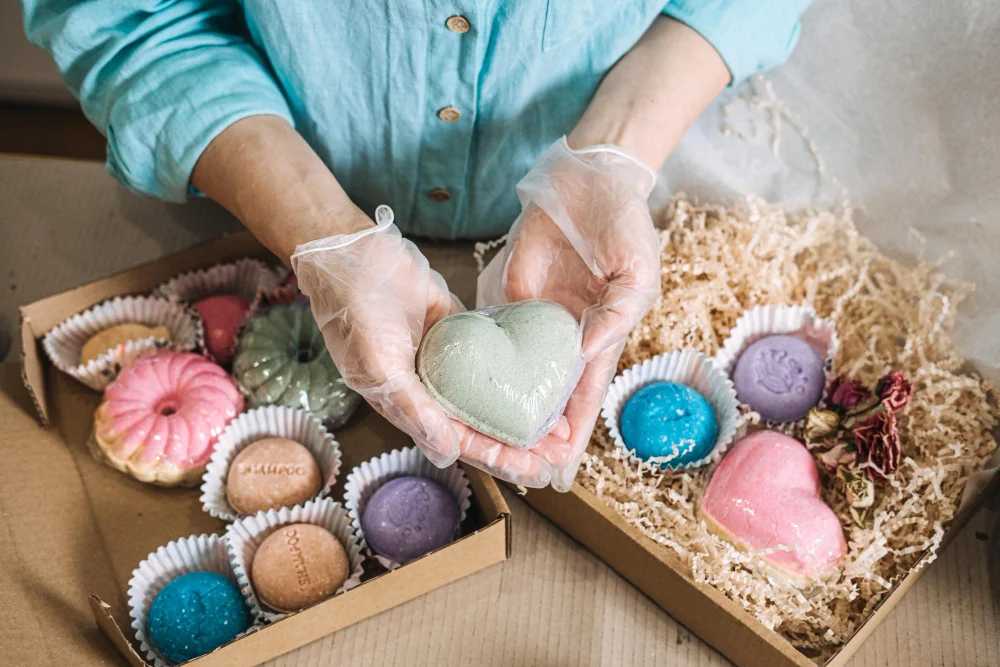 Person mit Handschuhen hält ein herzförmiges Badeprodukt über Geschenkboxen mit bunten handgefertigten Seifen und Badebomben