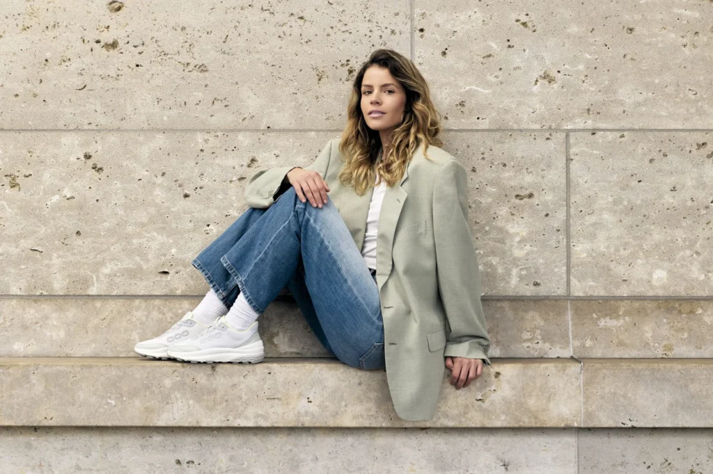 Frau in Jeans und Blazer mit weißen Sneakern vor einer Natursteinwand