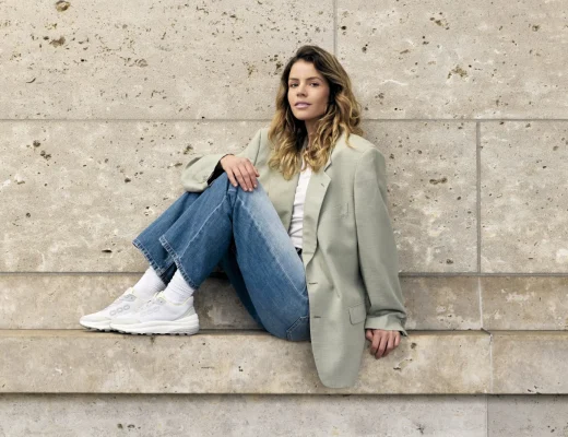 Frau in Jeans und Blazer mit weißen Sneakern vor einer Natursteinwand