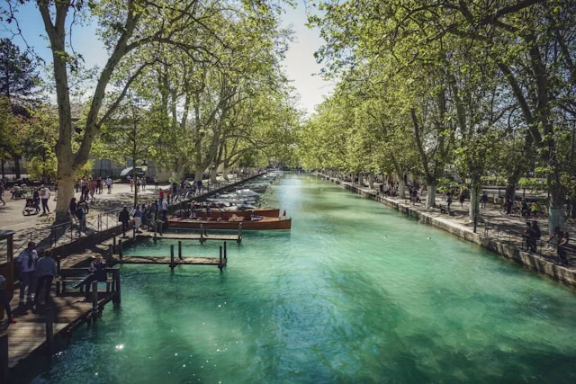 Türkisfarbener Kanal in Annecy mit Bäumen, Booten und Spaziergängern am Ufer