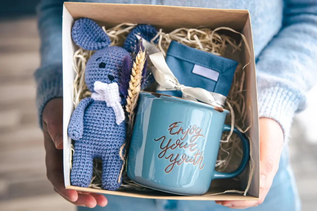 Hände halten eine Geschenkbox mit blauem Strickhasen, Tasse und Stoffbeutel in Strohfüllung