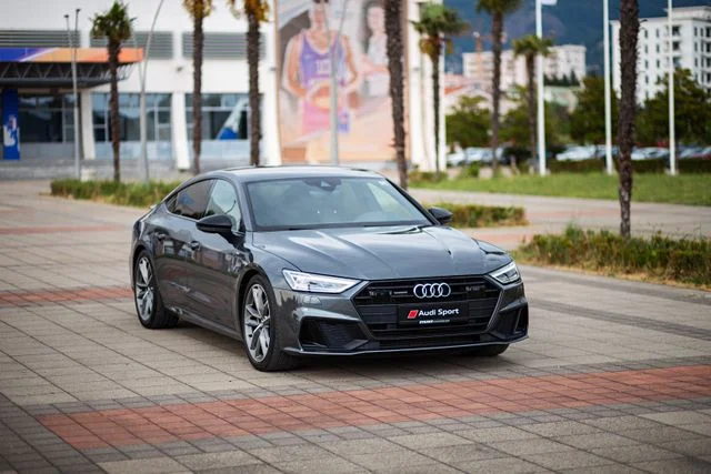 Ein grauer Audi A7 Sportsback ist auf einem urbanen Gehweg mit Palmen geparkt.